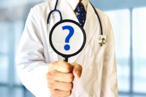 聴診器を首にかけ白衣を着た男性医師がクエスチョンマークを書いた虫眼鏡を持っているところ、患者の質問に答えるイメージ